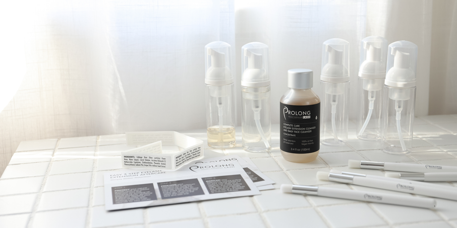  Prolong Lash cleanser products arranged on a white tiled surface with foaming bottles, a concentrate bottle, instruction cards, and cleansing brushes. 